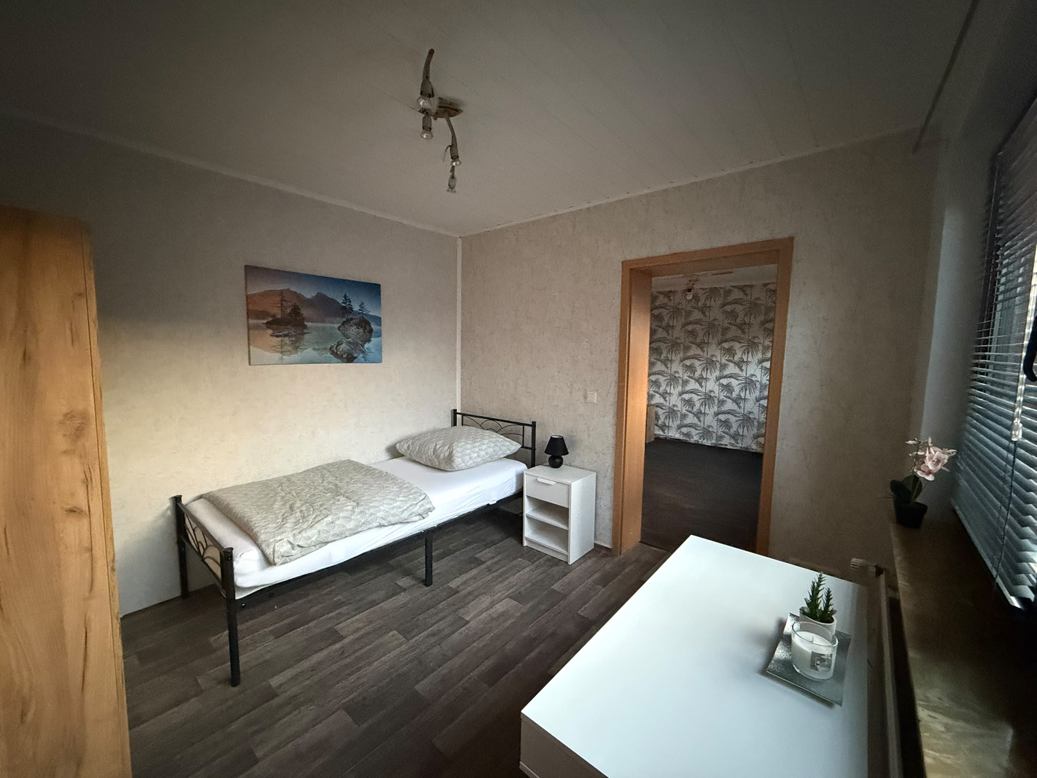 Möbliertes Zimmer mit Einzelbett, Nachttisch, weißem Tisch am Fenster und offener Tür zum angrenzenden Raum.