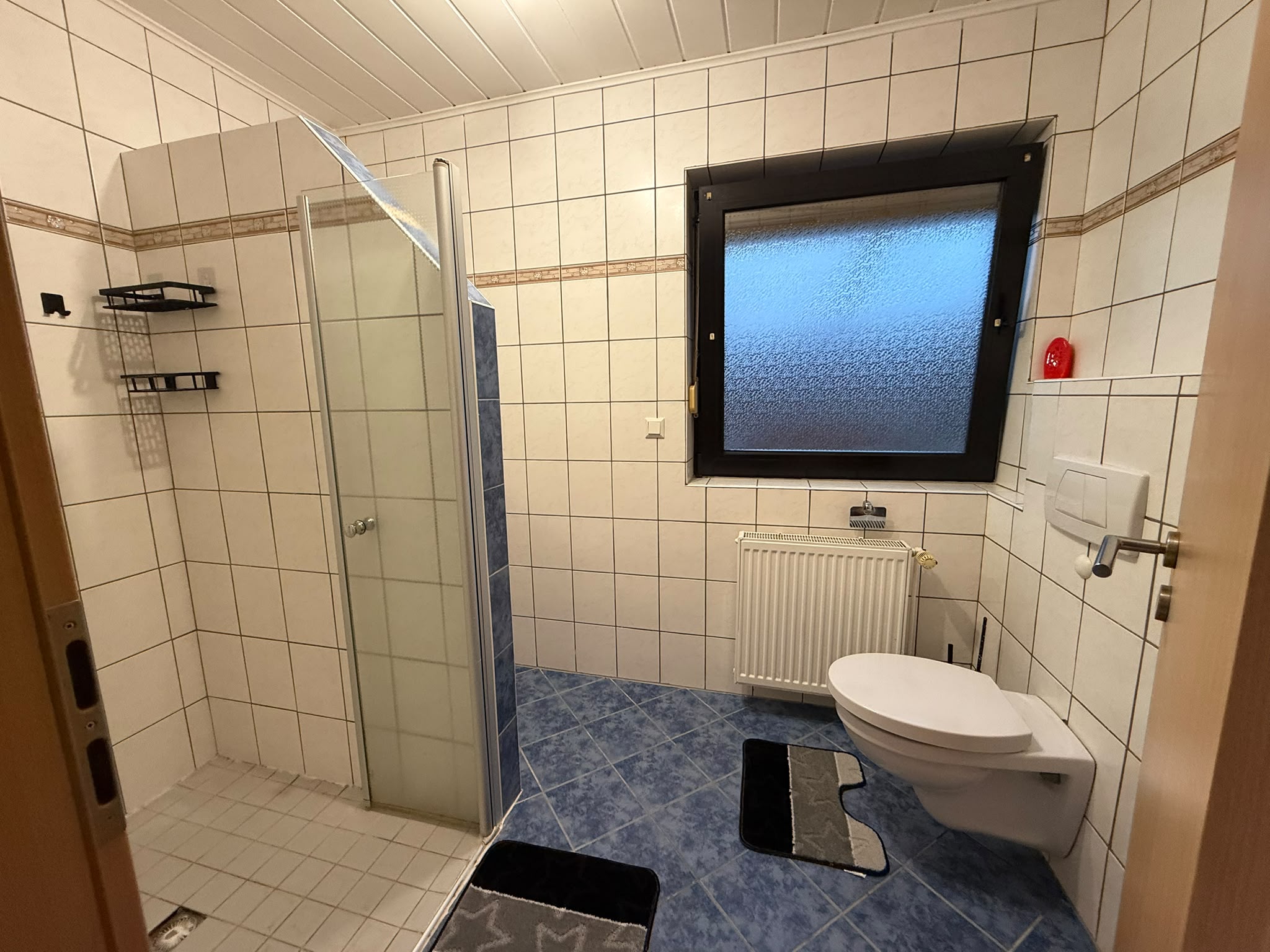 Badezimmer mit gefliester Dusche und Glasabtrennung, WC, Fenster und Heizkörper.